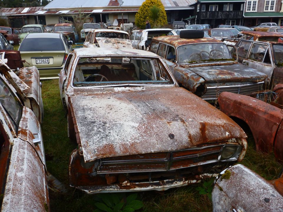 Front view of a heavily rusted car in a junkyard, with other vehicles visible