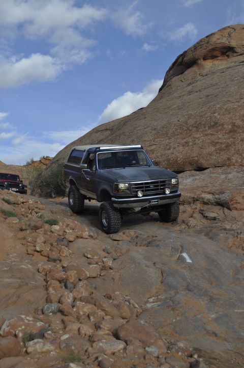1991 Ford Bronco - A dark gray Ford Bronco navigates a rocky off-road path under a cloudy sky, with another vehicle visible in the background.