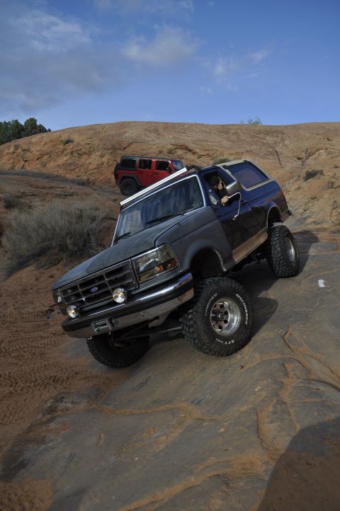1991 Ford Bronco - Two off-road vehicles navigate a rocky, desert terrain under a blue sky.