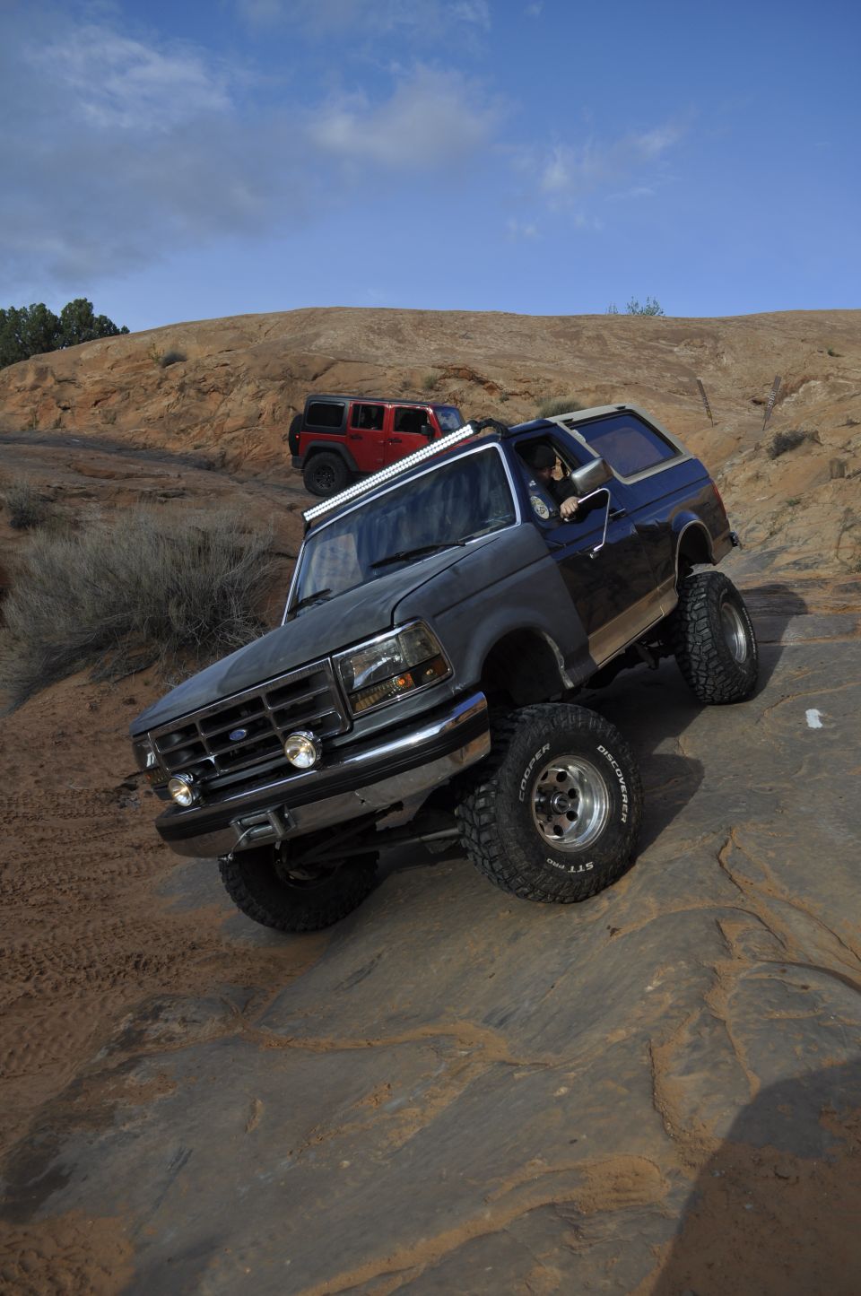 Dark gray Ford Bronco climbing rocks, red Jeep in background