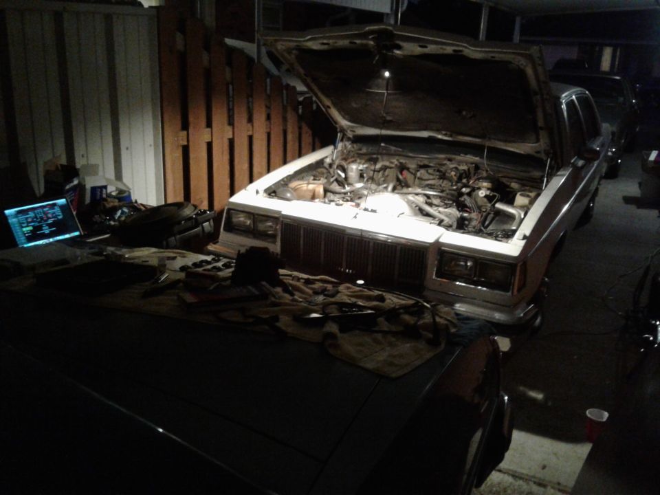 White 1980s sedan with hood open, engine exposed, laptop with diagnostics nearby in garage.