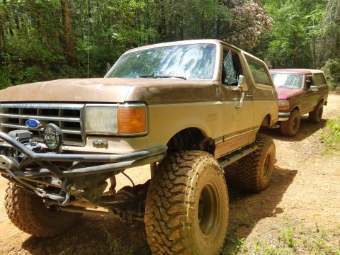 1988 Ford Bronco - A lifted, mud-covered tan 1988 Ford Bronco is parked on a dirt road in a wooded area, with a red Bronco following behind it.