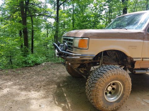 1988 Ford Bronco - A lifted, tan 1988 Ford Bronco with large off-road tires is parked on a dirt path in a wooded area. The front of the vehicle features a cus…