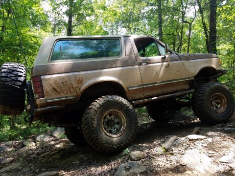 1988 Ford Bronco - A lifted, mud-splattered 1988 Ford Bronco is parked on a rocky, wooded trail. A spare tire is mounted on the rear driver's side.