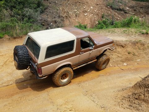 1988 Ford Bronco - A two-tone brown and cream 1988 Ford Bronco is shown from a rear-three-quarter angle, driving through a muddy trail with large off-road tir…
