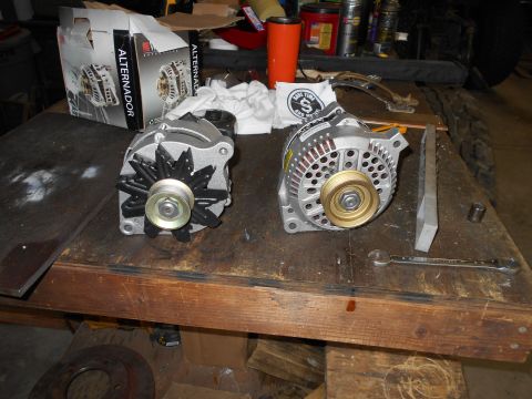 1992 Ford Bronco - Two automotive alternators are displayed on a wooden workbench. One alternator is in its original box, while the other is removed and ready…
