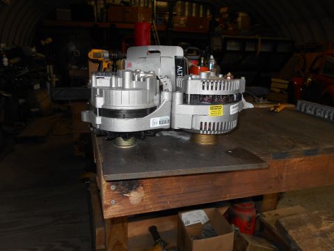 1992 Ford Bronco - Two large silver alternators are mounted on a metal plate on top of a wooden workbench in a garage. A yellow and black sticker on the right…