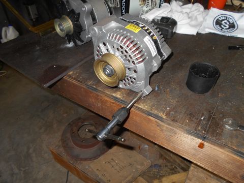 1992 Ford Bronco - An automotive alternator with a brass pulley is mounted on a wooden workbench next to a brake rotor and a tool. The alternator is silver wi…