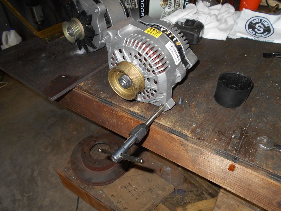 Silver alternator with brass pulley on workbench next to brake rotor and tool.
