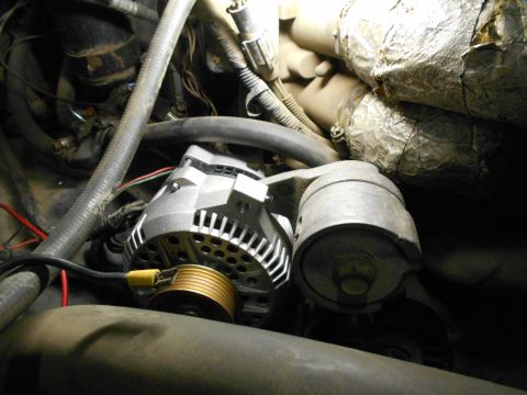 1992 Ford Bronco - Close-up view of an alternator and its mounting bracket in an engine bay, with various hoses and wiring visible. The alternator is silver w…