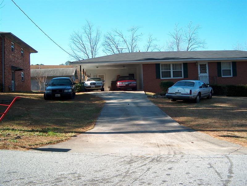 Four cars parked in front of a house and carport. One is a dark sedan, two are lighter sedans, and one is a red sedan