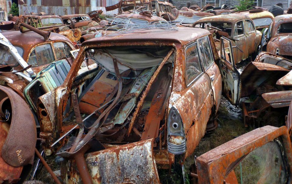 Junkyard scene with many heavily rusted cars piled together. Focus on the rear of a station wagon with its interior exposed