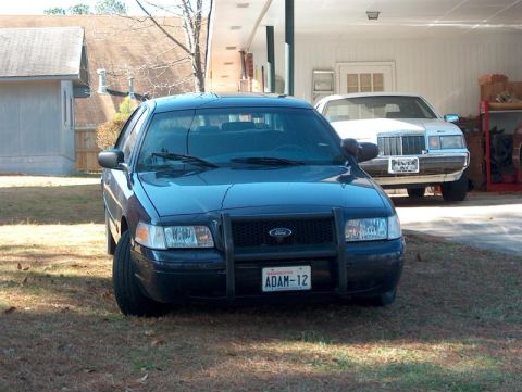 2001 Ford Crown Victoria - A black 2001 Ford Crown Victoria is parked in front of a white building, with another car behind it.