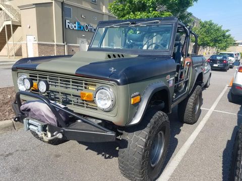 1972 Ford Bronco - A modified olive green and black 1972 Ford Bronco is parked in a lot in front of a FedEx Office building. The Bronco features large off-roa…