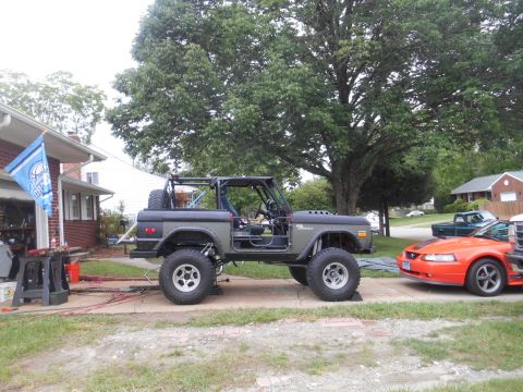 1972 Ford Bronco - A dark green 1972 Ford Bronco is parked on a concrete driveway next to a house. The Bronco has a roll cage, large off-road tires, and appea…