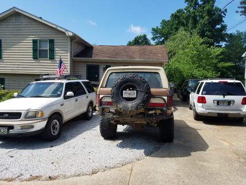 1988 Ford Bronco - A lifted, tan and brown 1980s Ford Bronco with a spare tire mounted on the rear is parked on a gravel driveway next to a white Ford Expedit…