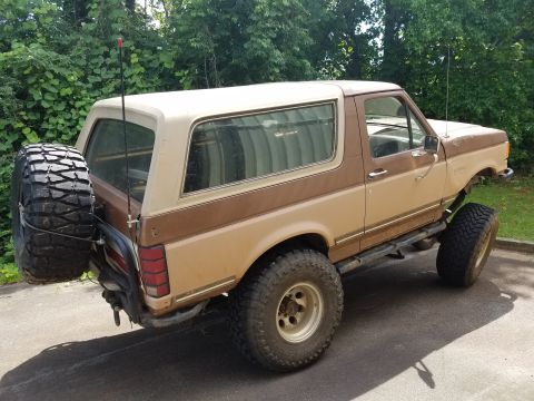 1988 Ford Bronco - The rear passenger side of a lifted, two-tone brown and tan 1980s Ford Bronco is shown parked on asphalt. A large spare tire is mounted to…