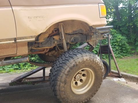 1988 Ford Bronco - The side view of a tan Ford Bronco is shown lifted on a jack stand with a large off-road tire prominently displayed. The vehicle's suspensi…