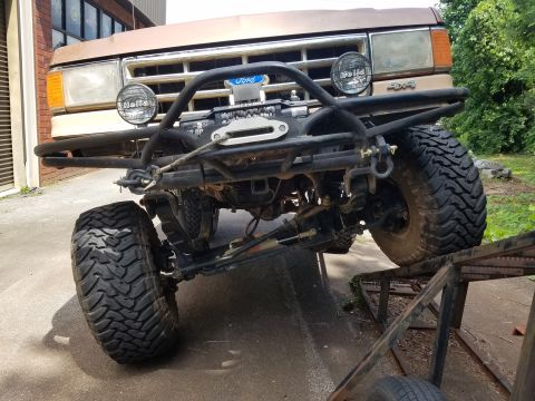 1988 Ford Bronco - The front end of a lifted, brown 1988 Ford Bronco is shown angled upwards on a ramp. It features a custom black metal bumper with a winch,…