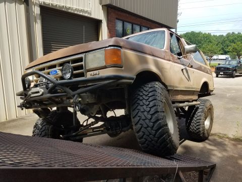 1988 Ford Bronco - A heavily modified, lifted 1980s Ford Bronco is parked on a metal ramp with a custom front bumper and winch. The vehicle is a two-tone brow…