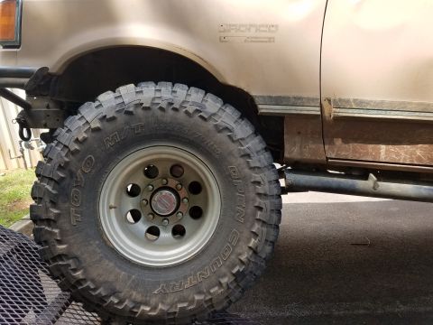 1988 Ford Bronco - The front driver's side wheel and fender of a tan Ford Bronco are shown. The vehicle has a large, aggressive off-road tire and a visible af…