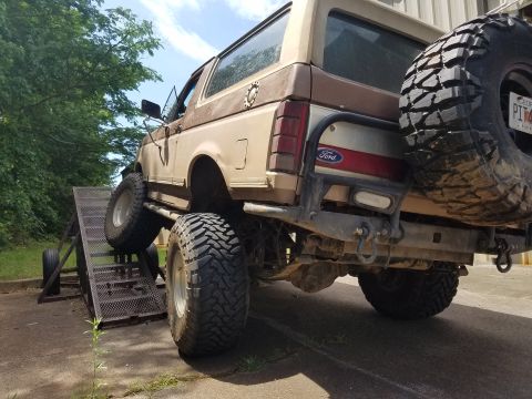 1988 Ford Bronco - The rear driver's side of a lifted, two-tone brown 1980s Ford Bronco is shown on a metal ramp. The vehicle has large off-road tires and a s…