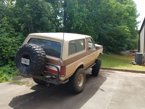 1988 Ford Bronco - The rear passenger side of a lifted, two-tone brown and tan Ford Bronco is shown parked on asphalt. A large spare tire is mounted to the re…