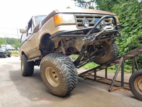 1988 Ford Bronco - A lifted, tan and brown Ford Bronco is positioned on metal ramps, showcasing its large off-road tires and custom front bumper with a winch.…