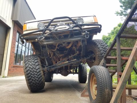 1988 Ford Bronco - The underside of a lifted 1988 Ford Bronco with large off-road tires is shown, with a metal ramp structure to its right. The vehicle is par…