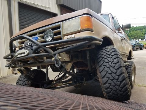 1988 Ford Bronco - The front quarter view of a lifted, tan 1980s Ford Bronco with a custom heavy-duty front bumper and winch. The vehicle is parked outdoors n…