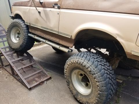 1988 Ford Bronco - A lifted, tan 1988 Ford Bronco is partially on a metal ramp. The vehicle has large, aggressive off-road tires and prominent side steps.