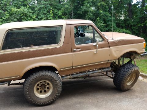 1988 Ford Bronco - The side profile of a lifted, two-tone brown 1988 Ford Bronco is shown, featuring large off-road tires and custom side steps. The vehicle i…