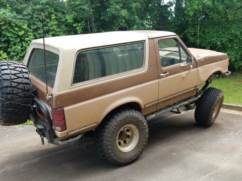 1988 Ford Bronco - A lifted, two-tone brown and tan 1980s Ford Bronco is parked outdoors with a spare tire mounted on the rear. The vehicle has large off-road…