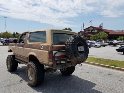 1988 Ford Bronco - A lifted, two-tone brown 1980s Ford Bronco is parked in a parking lot in front of a Bass Pro Shops store. The vehicle has large off-road ti…