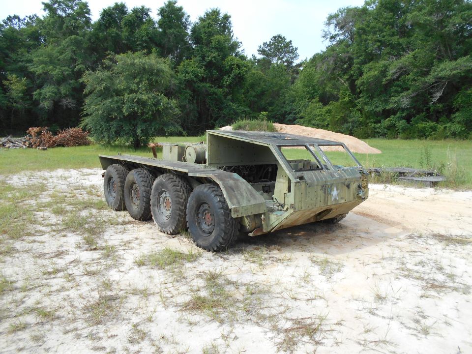 Olive drab six-wheeled amphibious military vehicle with large tires in a sandy, grassy area.