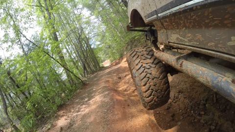 1988 Ford Bronco - A close-up, low-angle shot of the muddy tire and side of a tan off-road vehicle driving on a dirt trail through a forest.