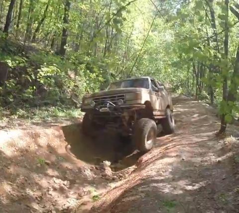 1988 Ford Bronco - A lifted, tan 1988 Ford Bronco is navigating a muddy, rutted trail through a dense forest.