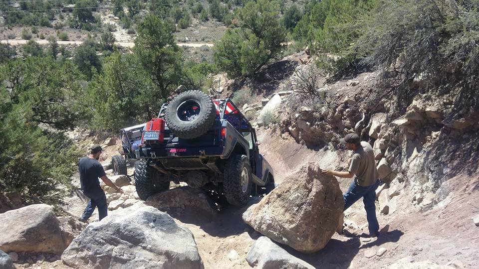 Modified dark Ford Bronco climbing a rocky off-road trail, with two men observing.