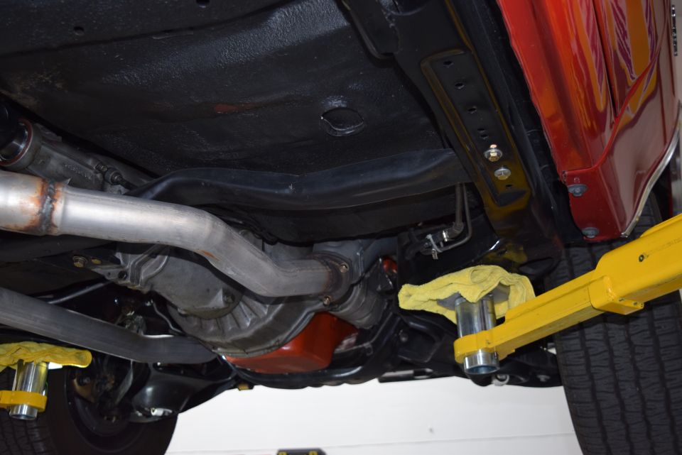 Underside of a classic car on a yellow lift, showing exhaust, drive train, and red oil pan.