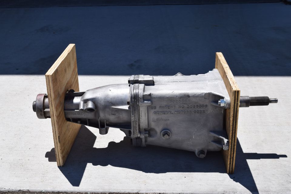 Silver manual transmission with General Motors Corp. patent information, mounted on plywood.