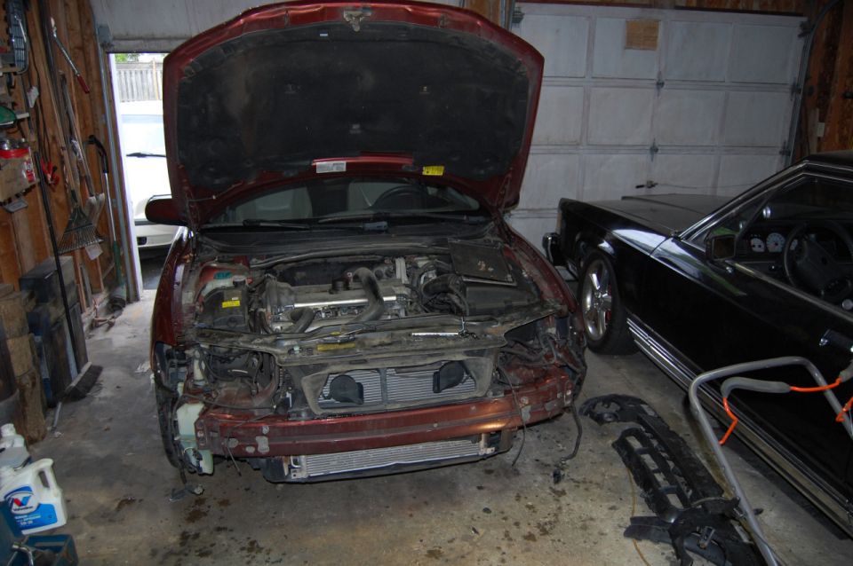 Maroon sedan with hood open, front bumper removed, parked in garage next to black sedan.