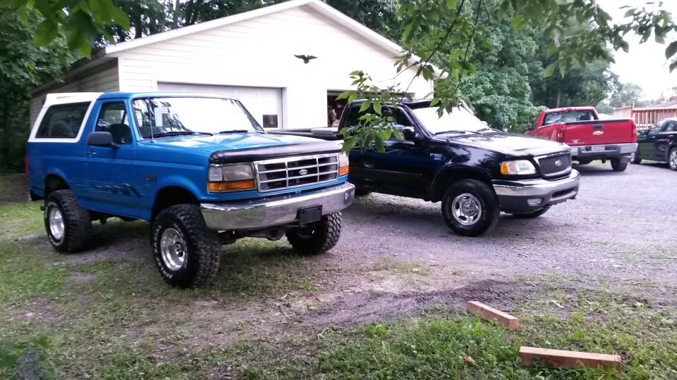 Lifted blue 1995 Ford Bronco and black Ford F-150 pickup trucks parked in front of a garage.