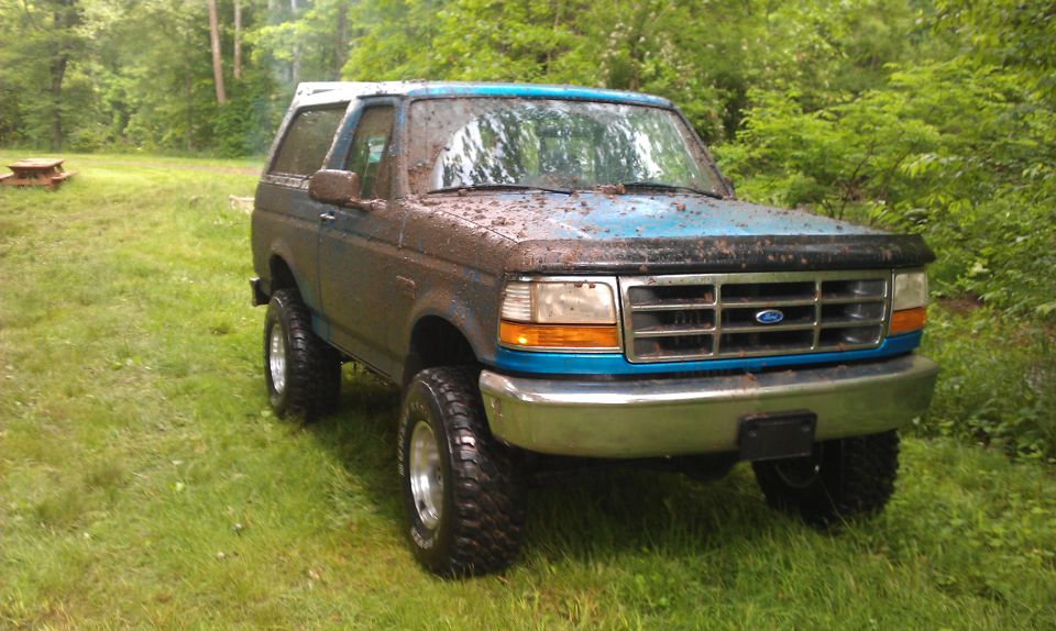 Muddy blue 1995 Ford Bronco parked in a grassy area with trees in the background.