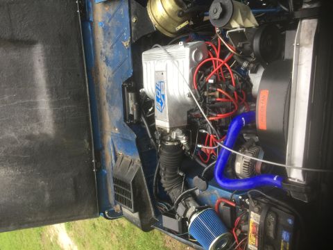 1974 Ford Bronco - The engine bay of a blue 1974 Ford Bronco shows a V8 engine with a polished aluminum intake manifold and red spark plug wires. A blue silic…