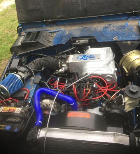 1974 Ford Bronco - The engine bay of a blue 1974 Ford Bronco shows a modified 5.0L V8 engine with a blue air filter and red spark plug wires. The hood is open…