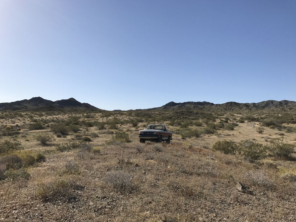 Light-colored Ford truck parked in a desert clearing with brush and mountains in the background