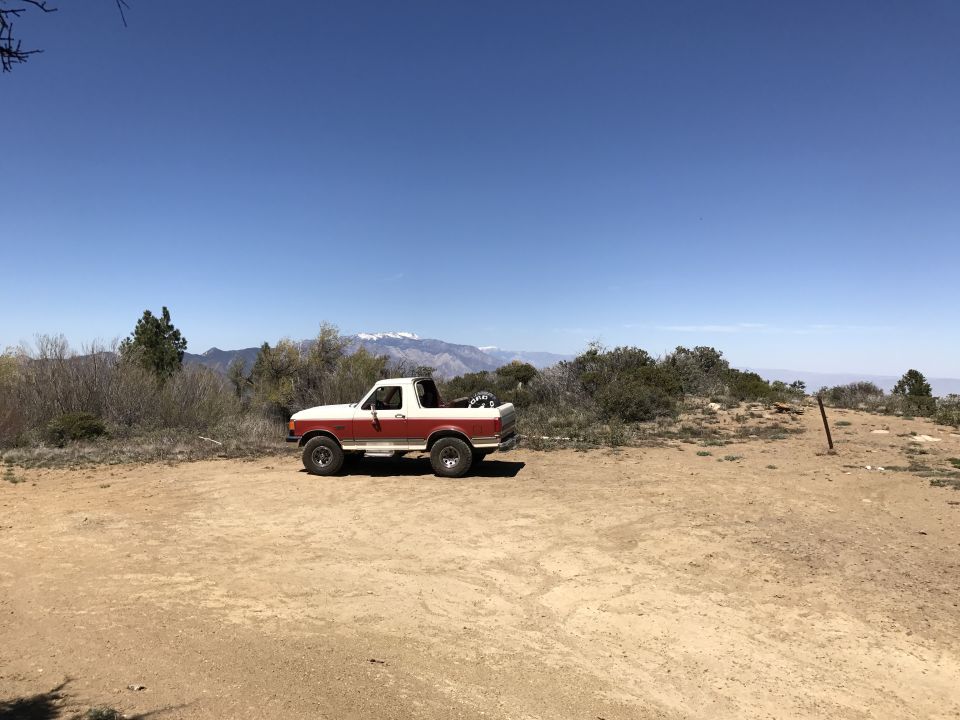 White and red 1987 Ford Bronco with top removed, parked on a dirt clearing with mountains behind