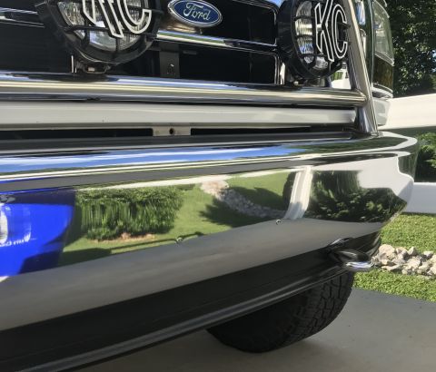 1995 Ford Bronco - Close-up front view of a chrome bumper on a Ford Bronco, reflecting a grassy yard and trees. A tow hook is visible on the right side of the…
