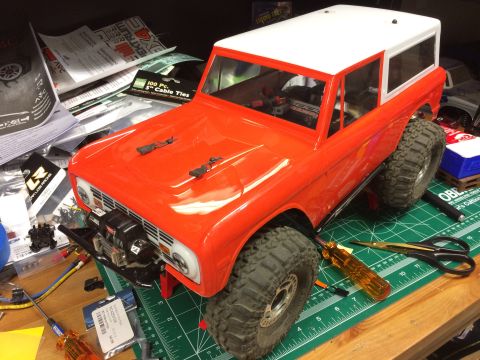 1989 Ford Bronco - An orange RC car body, resembling a classic SUV, sits on a workbench with tools and hobby supplies.