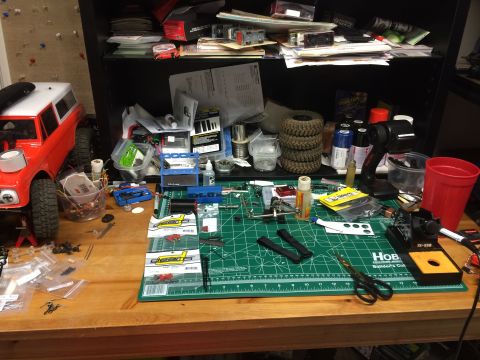 1989 Ford Bronco - RC car parts and tools are spread across a workbench next to a red and white RC vehicle.